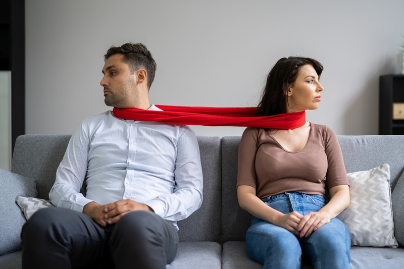 man and woman tied together by a red scarf sitting on a couch