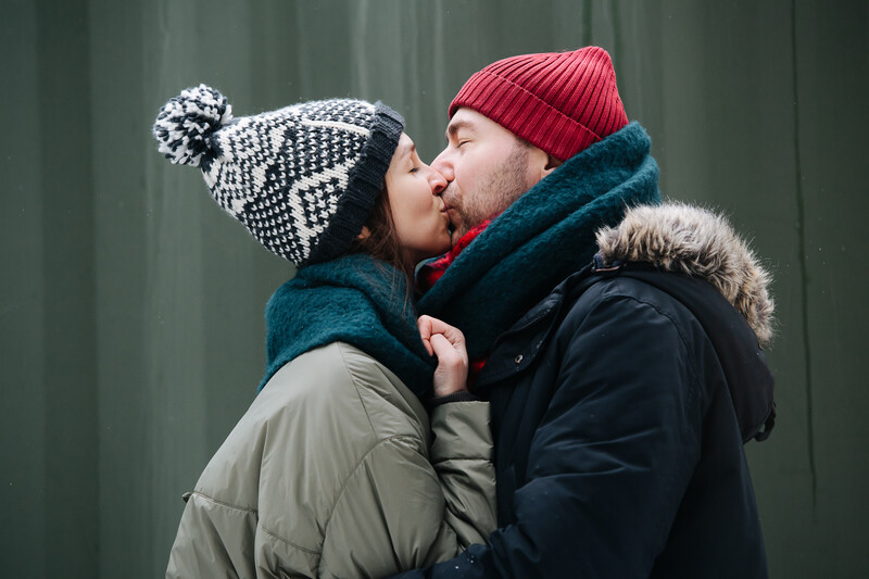 couple is hats and scarfs kissing in front of a barn